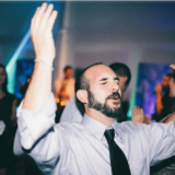 Man dancing with raised arms at lively Boston wedding celebration under colorful lights.