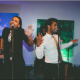 Two male singers performing passionately with a keyboardist at a vibrant Boston art venue.
