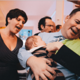 Father joyfully holding baby amid family celebration and laughter indoors.