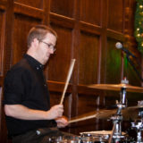 Focused drummer performing at festive holiday event in warm, wood-paneled venue with glowing wreath.