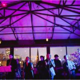 Live band performing at elegant glass-roof rooftop venue with city skyline and vibrant lighting.