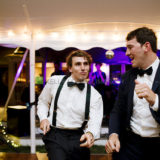 Two men joyfully dancing at an elegant wedding reception under glowing string lights.