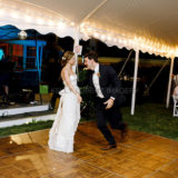 Bride and groom dancing joyfully beneath glowing string lights at elegant outdoor wedding reception.