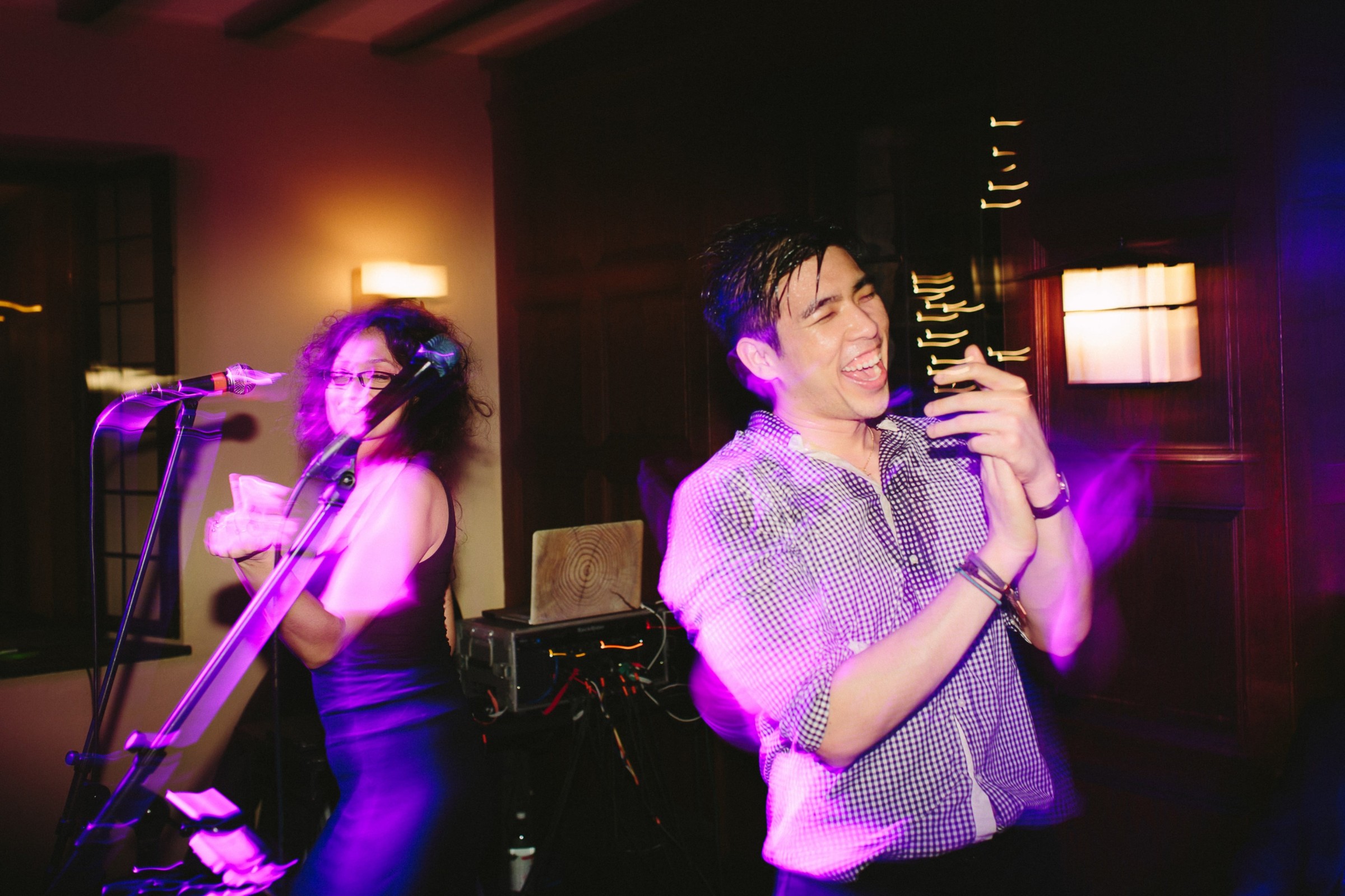 Joyful couple dancing and playing violin under vibrant pink and purple lights at indoor celebration.