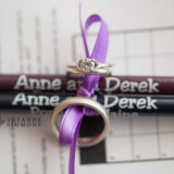 Wedding rings tied with purple ribbon beside personalized pencils on a crossword puzzle.