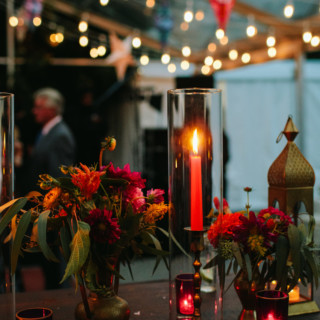 Romantic outdoor wedding table with red candles, brass vases, and glowing string lights at dusk.