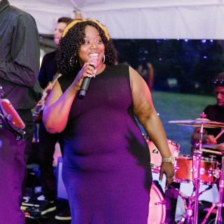 Joyful female singer performing live with band under glowing string lights at outdoor evening event.