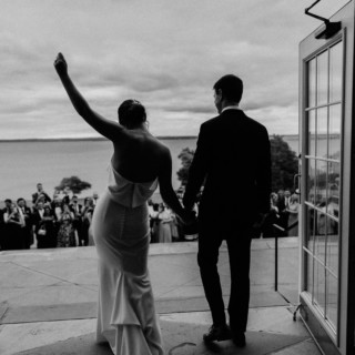 Bride and groom celebrate at Aldrich Mansion overlooking waterfront under dramatic cloudy sky.
