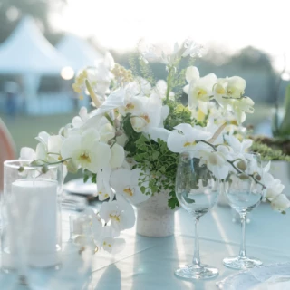 Elegant outdoor wedding table with white orchid centerpiece, glassware, and candle in soft sunlight.
