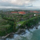 Aerial view of a luxury cliffside resort in Bali with ocean waves and lush greenery.