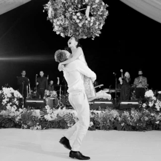 Groom lifting laughing bride during joyful black-and-white wedding dance under floral canopy in Bali.