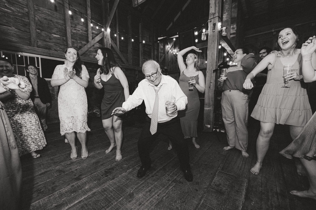 Older man dancing joyfully at rustic barn wedding surrounded by laughing guests and warm string lights.