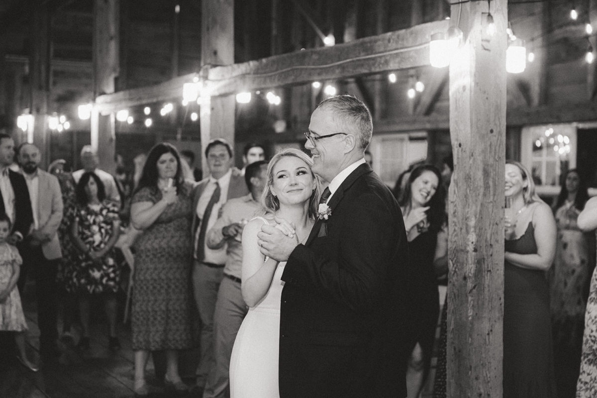 Bride and father share emotional dance at rustic barn wedding with warm string lights.