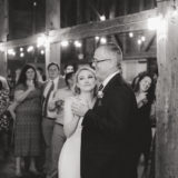 Bride and father share emotional dance at rustic barn wedding with warm string lights.