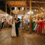 Bride and father share emotional dance at rustic barn wedding surrounded by warm lights and guests.