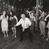 Older man dancing joyfully at rustic barn wedding surrounded by laughing guests and warm string lights.