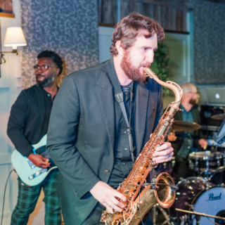 Jazz trio performing live in cozy indoor venue with saxophonist, guitarist, and drummer.