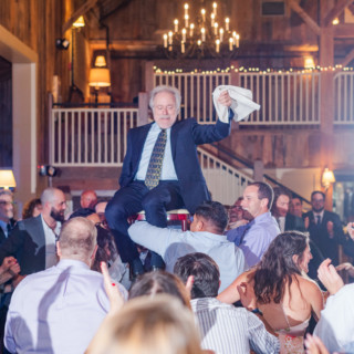 Guests lift an older man during a joyful Hora dance at a rustic Gibbet Hill wedding.