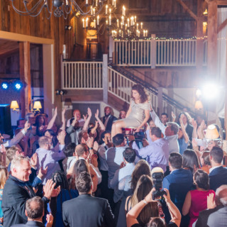 Bride lifted on chair during joyful rustic barn wedding celebration in Groton, Massachusetts.