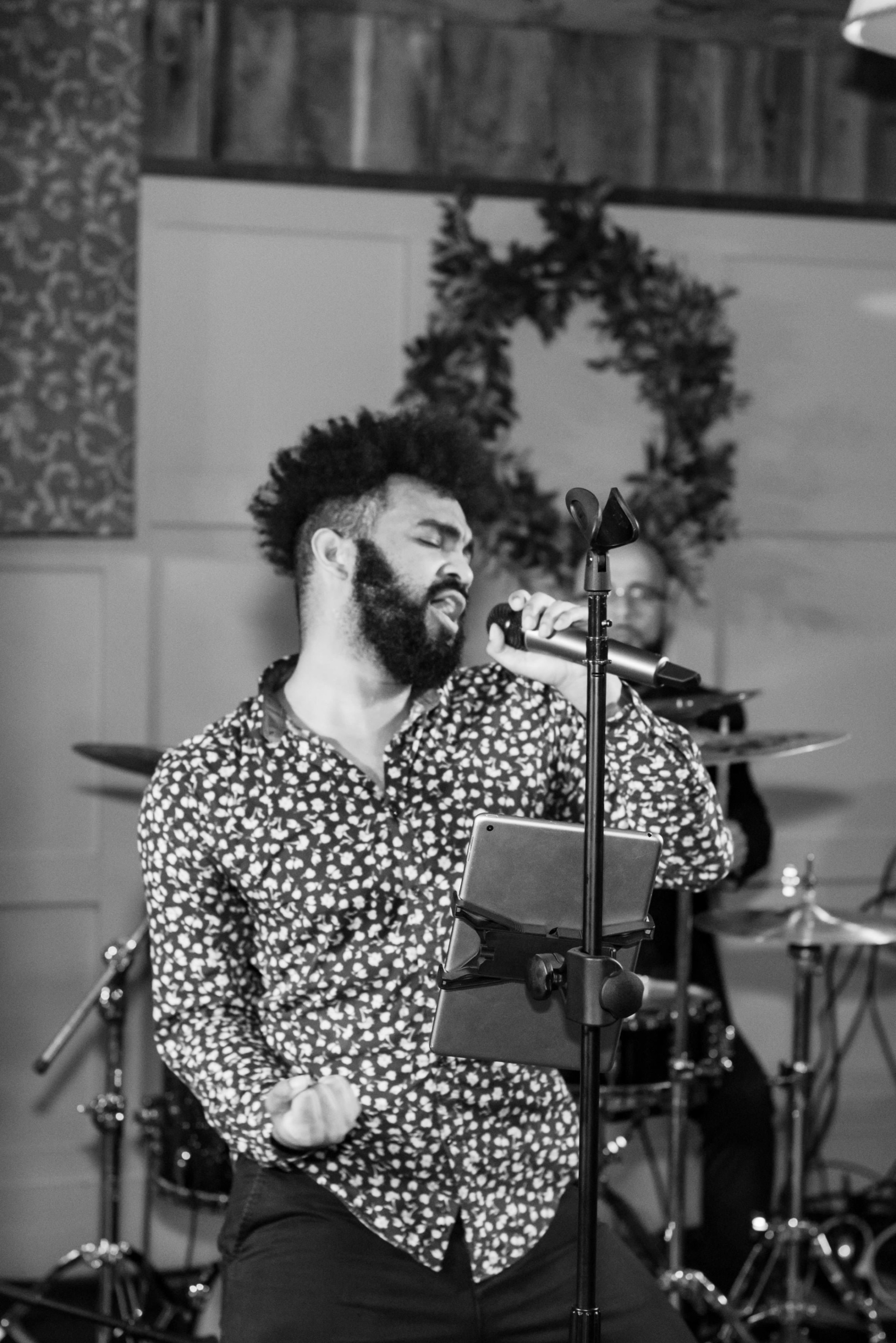 Emotional black and white photo of soulful male singer performing live with drummer in background.
