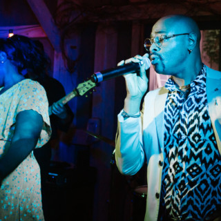 Singer performing under blue and purple lights with dancer and bassist at lively indoor music event.