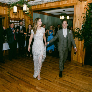 Bride and groom enter rustic Maine wedding reception surrounded by cheering guests and warm ambient lighting.