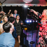Energetic trombone player performing for joyful wedding guests under glowing tent lights in Marblehead.
