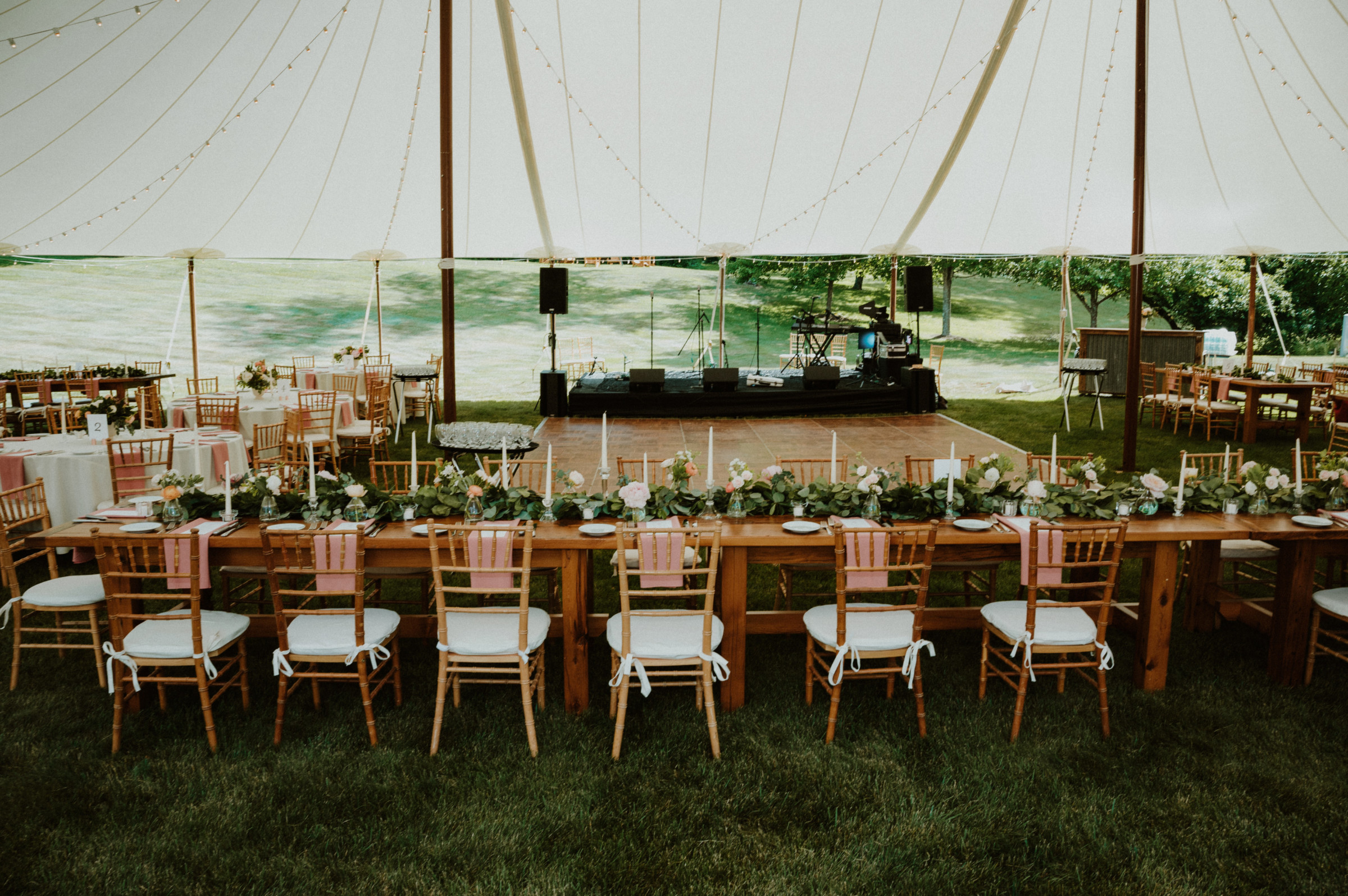 Elegant outdoor wedding reception under white tent with wooden tables, string lights, and floral décor.