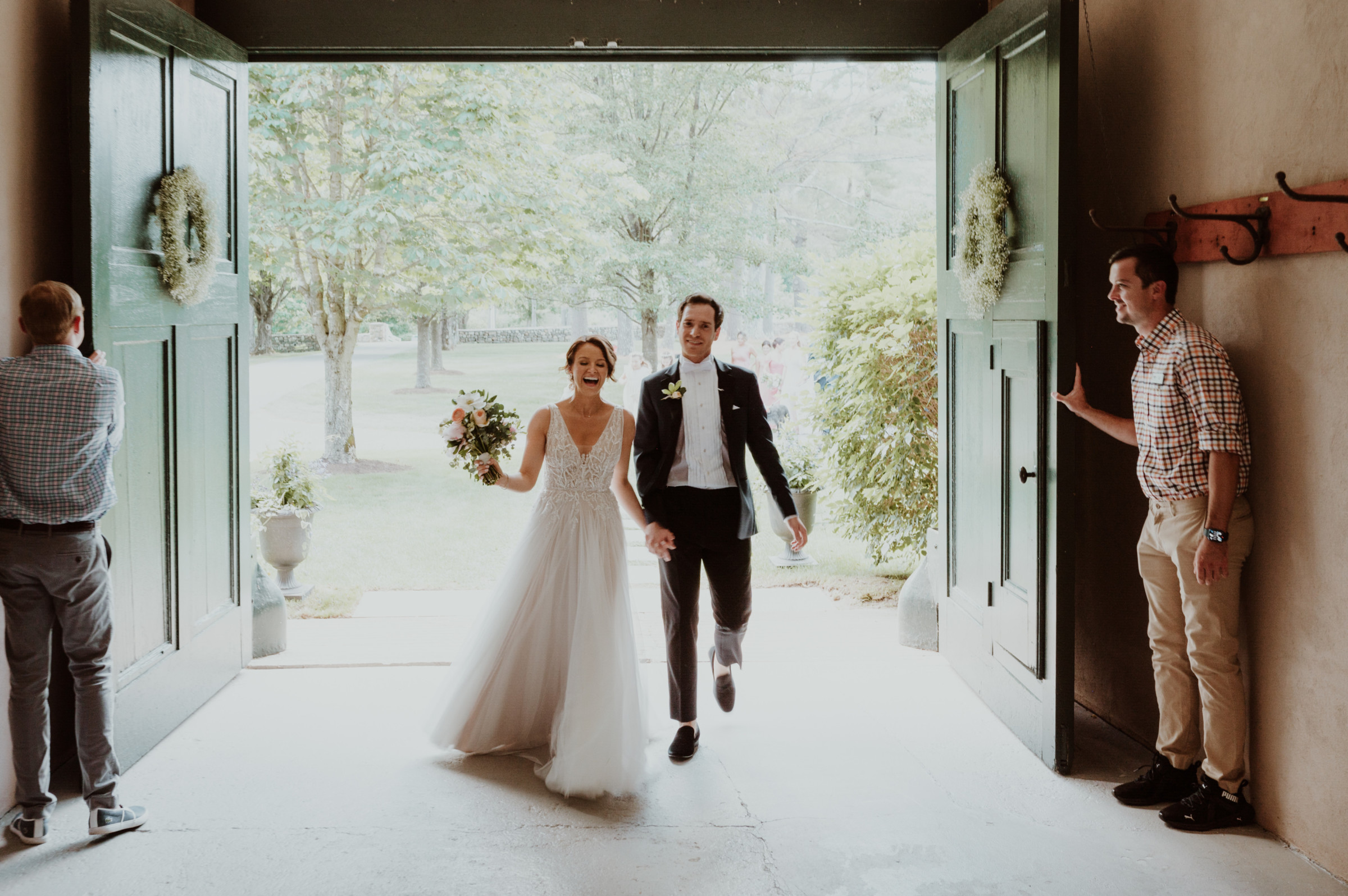 Smiling bride and groom walk through green doors into a sunlit garden wedding celebration.