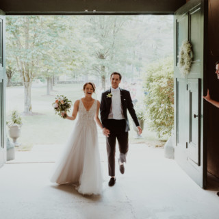 Smiling bride and groom walk through green doors into a sunlit garden wedding celebration.