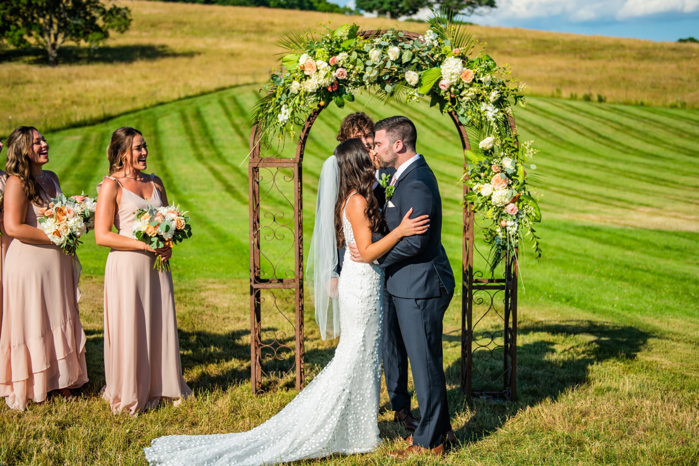 Bride and groom kiss under floral arch during romantic outdoor wedding at The Barn at Gibbet Hill.
