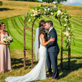 Bride and groom kiss under floral arch during romantic outdoor wedding at The Barn at Gibbet Hill.
