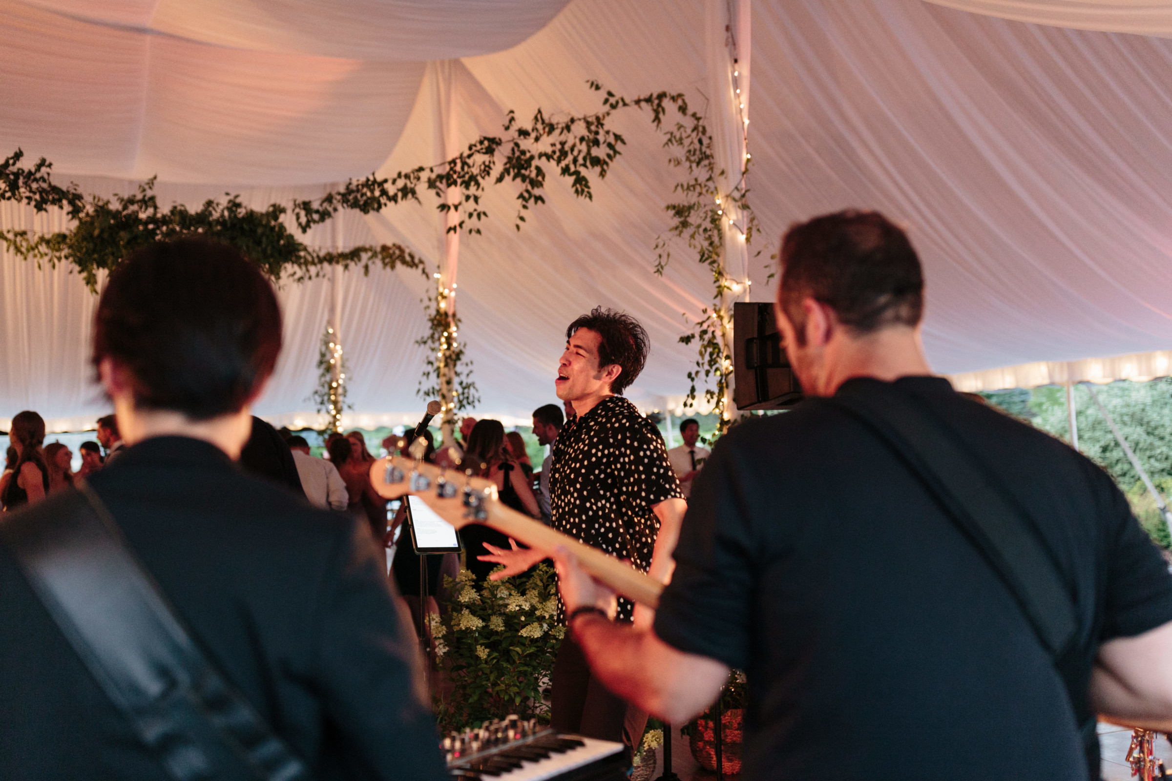Live band performing at elegant wedding reception under white tent with lights and greenery.