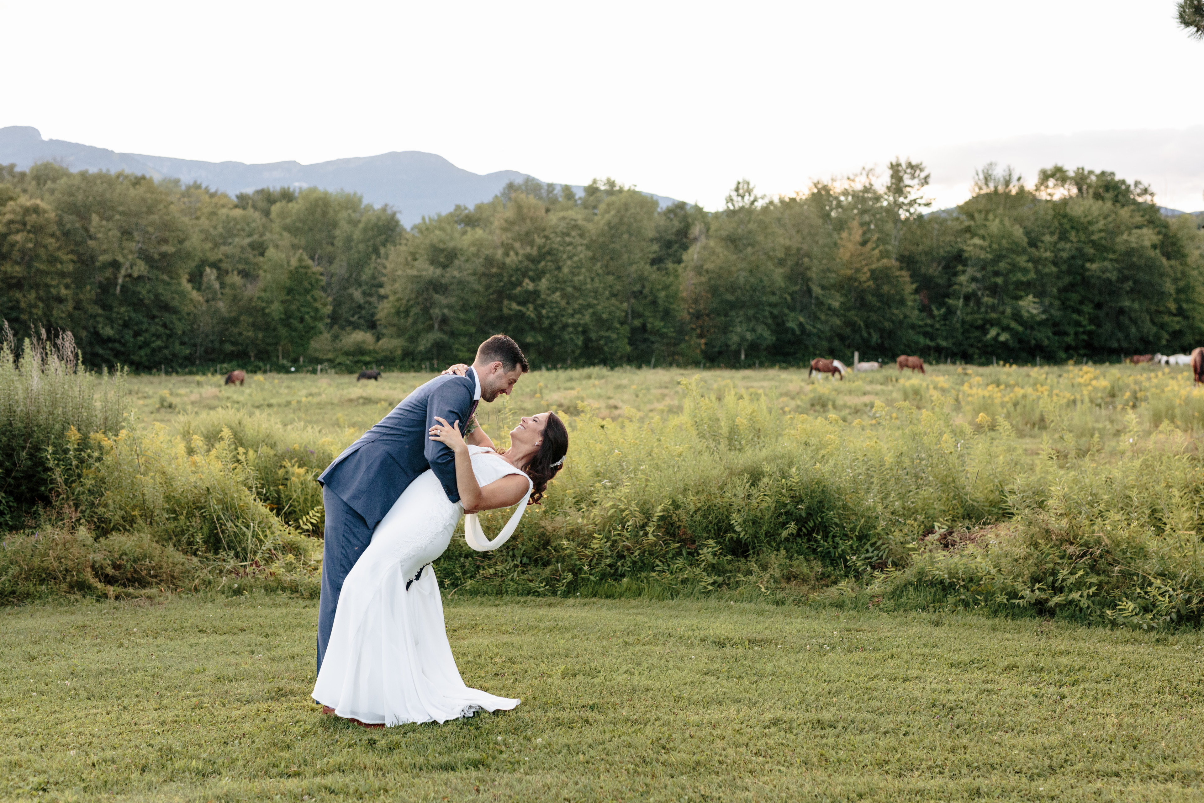 Bride and groom share a romantic dip in a scenic Vermont field with mountains and horses.