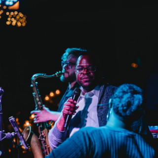 Vocalist and saxophonist performing under moody blue and purple stage lights at an intimate venue.