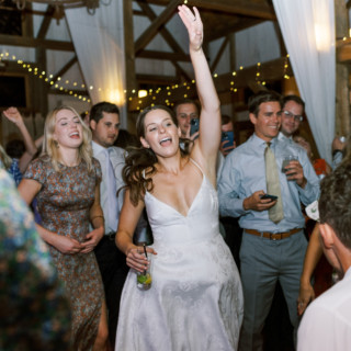 Bride dancing joyfully at rustic barn wedding surrounded by guests under warm string lights.