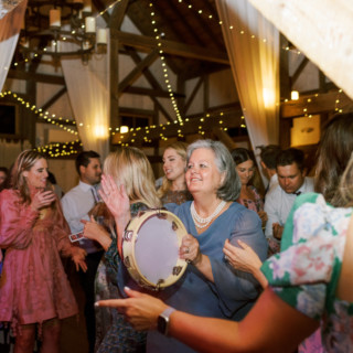 Elderly woman dancing with tambourine at rustic barn wedding reception under warm string lights.