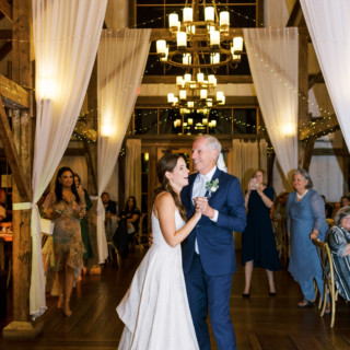 Father-daughter dance at rustic barn wedding surrounded by warm lights and joyful guests.