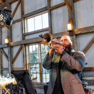 Trumpet player performing at rustic wedding reception with warm lighting and lively celebration atmosphere.