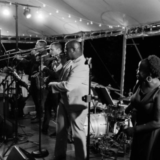 Black and white photo of a lively jazz band performing under wedding tent with string lights.