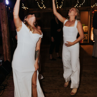 Two women dancing joyfully in white at a rustic barn wedding celebration in Maine.