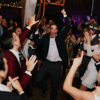 Man lifts chair as guests cheer during lively Cunningham Farm wedding reception in Maine.