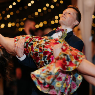 Groom dipping bride in floral dress during romantic dance under golden string lights at wedding.