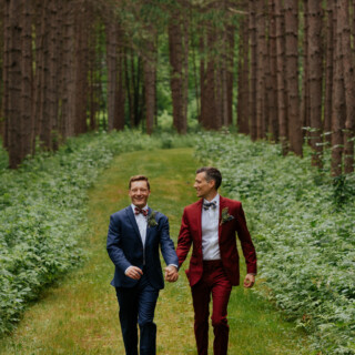 Two grooms walk hand in hand through a pine forest wedding path in Maine.