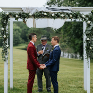 Two grooms exchange vows under a white floral arch at Cunningham Farm in Maine.