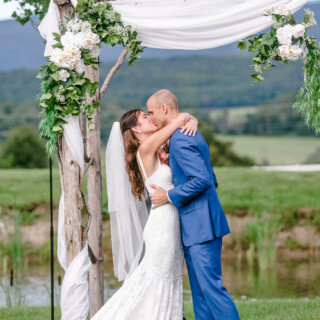 Newlyweds share a romantic first kiss under a rustic floral arch in Vermont countryside.