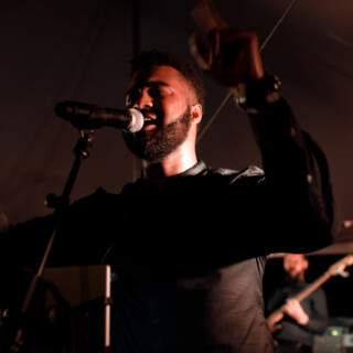 Bearded male singer performing passionately under warm stage lights with bassist in the background.