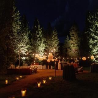 Romantic nighttime forest wedding with lanterns, string lights, and guests celebrating under tall pine trees.