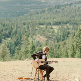 Elegant guitarist performing outdoors amid Montana mountain scenery and serene forest backdrop.