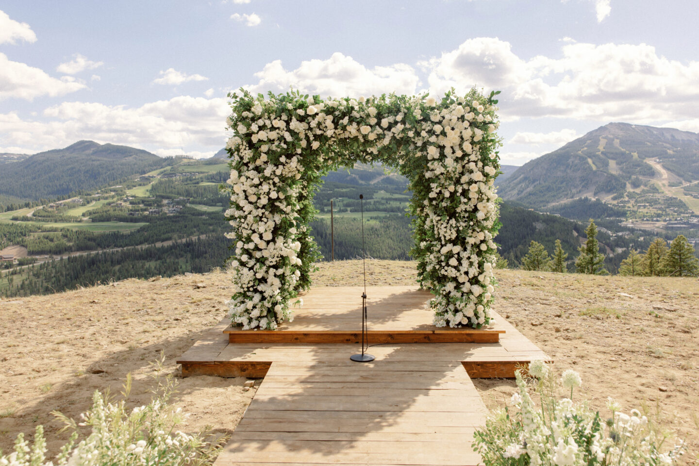 Scenic mountaintop wedding ceremony with white floral arch and panoramic mountain views in Big Sky Montana.